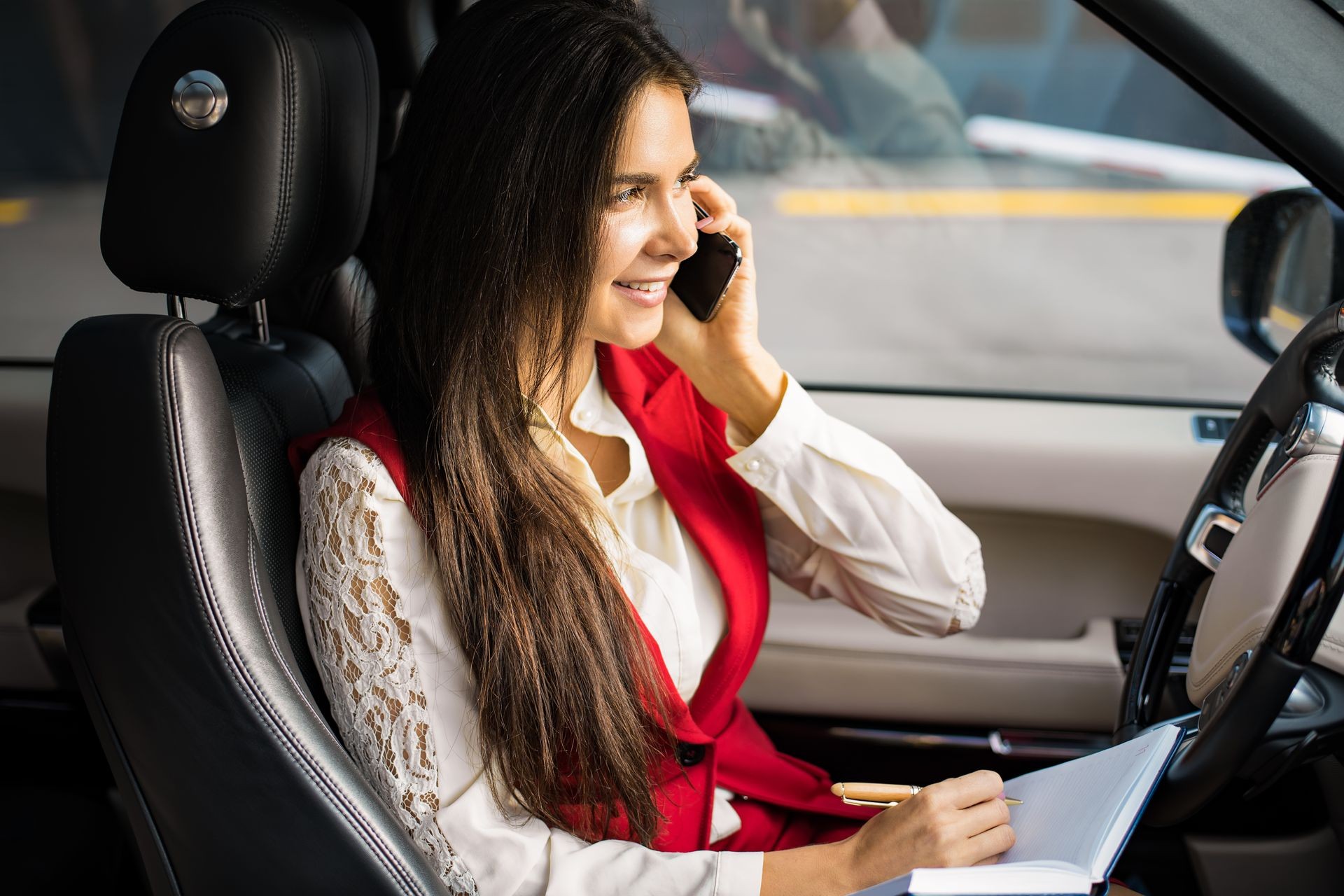 Smiling woman skilled entrepreneur having pleasant smartphone conversation and using textbook for notary, sitting in luxury automobile during break at job. Cheerful female executive director phone 